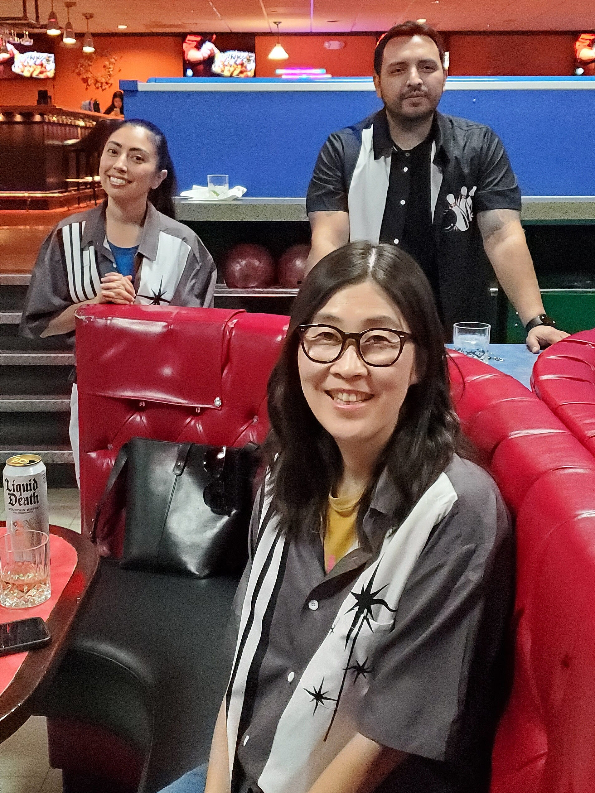 Three people in bowling shirts relaxing in a red booth, smiling at the camera.